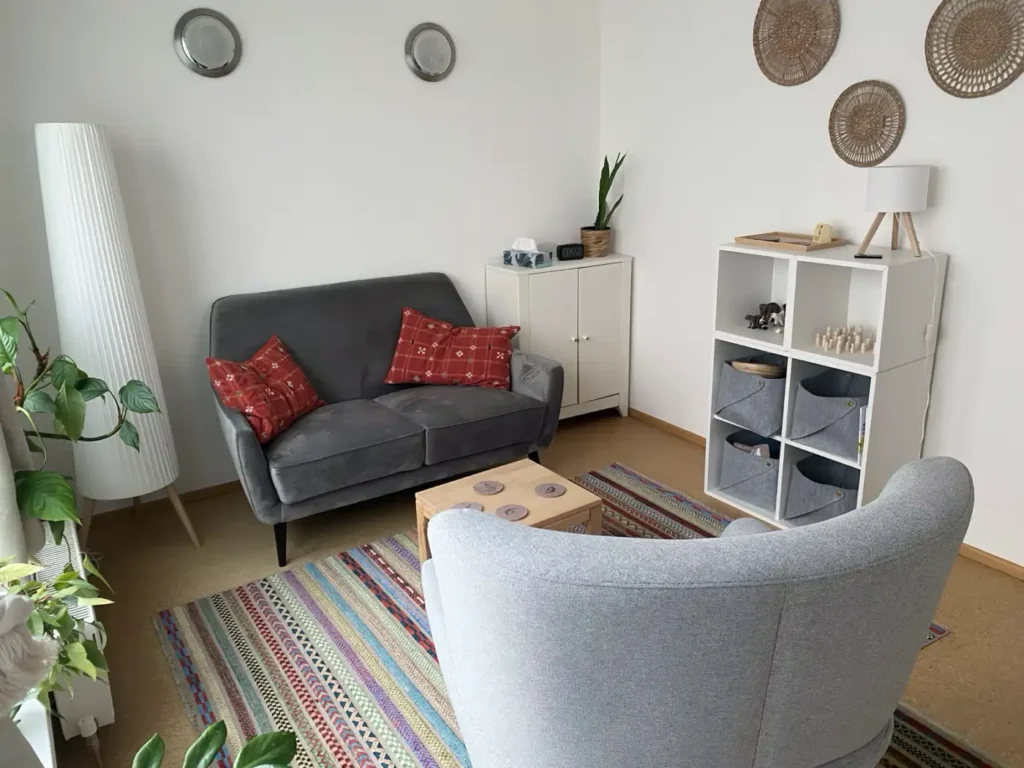 Heller Sitzbereich mit dunkelgrauem Sofa, hellgrauem Sessel, buntem Teppich, kleinem Holztisch und weißen Regalen